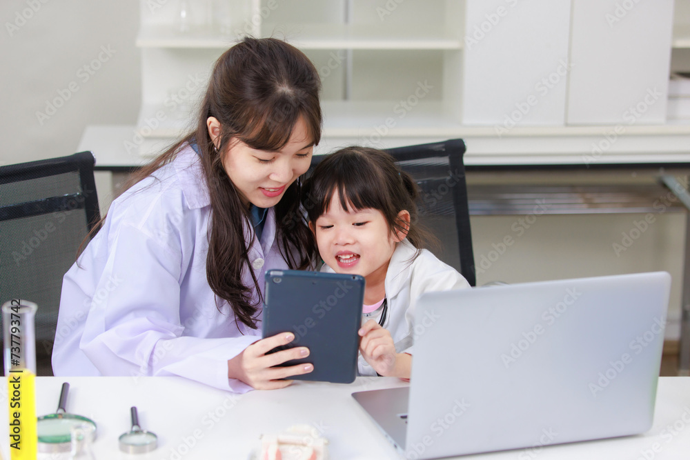 Asian woman scientist and little children girl reading laptop and tablet computer for data learning at chemical laboratory study room. Education research and development concept learning for kids. Asian woman scientist and little children girl reading laptop and tablet computer for data learning at chemical laboratory study room. Education research and development concept learning for kids.