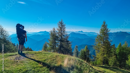 Man with baby carrier looking at magical mountain peaks of Karawanks and Julian Alps seen from Goldeck, Latschur group, Gailtal Alps, Carintha, Austria. Mystical atmosphere in Austrian Alps in summer