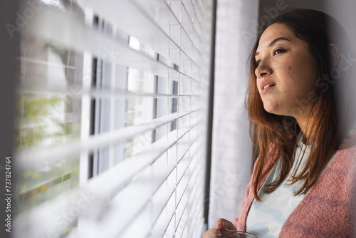 Young biracial plus size woman gazes out a window at home, with copy space photo