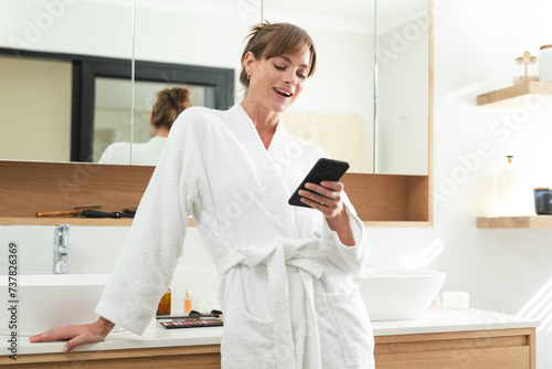 Caucasian middle-aged woman in a white robe enjoys her phone at home