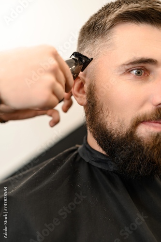 Barber shaves the contour of the oval line with a clipper on the client head. A man with a beard gets a haircut in a barbershop chair.