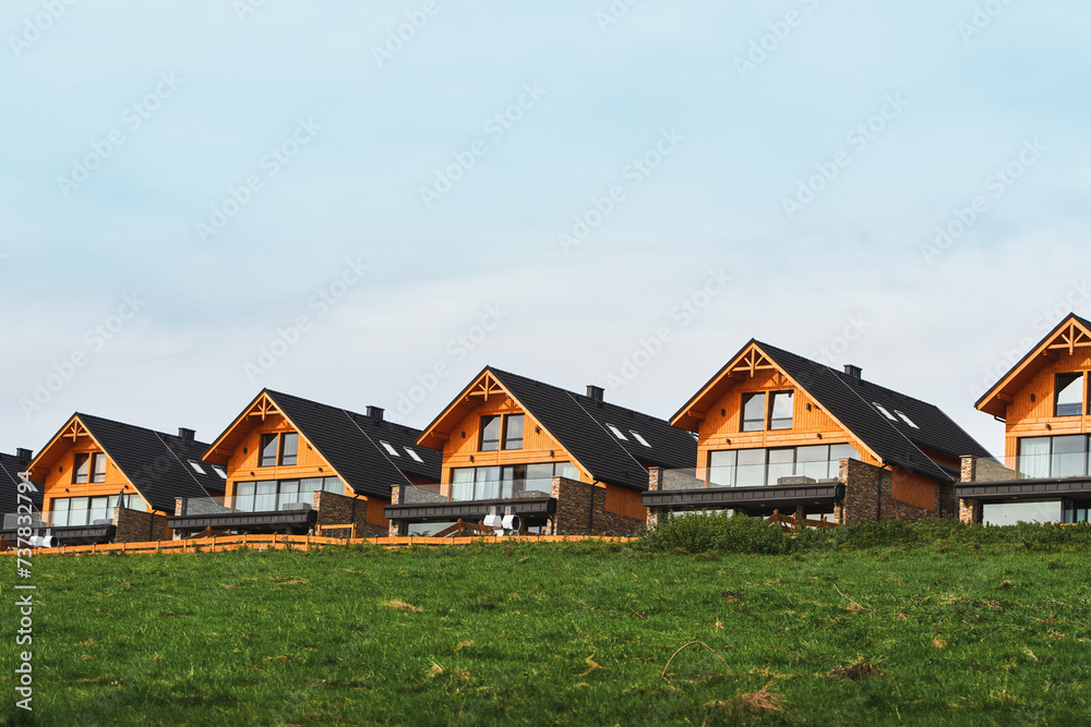 Wooden houses with modern design and architecture on a grassy hill overlooking the fields and the sky.