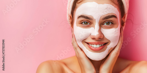 Smiling young woman applying clay mask on her face on pink background. Happy model cleansing beauty product  cream or scrub on cheeks. Cosmetic and skincare daily routine. Facial mask. Copy space
