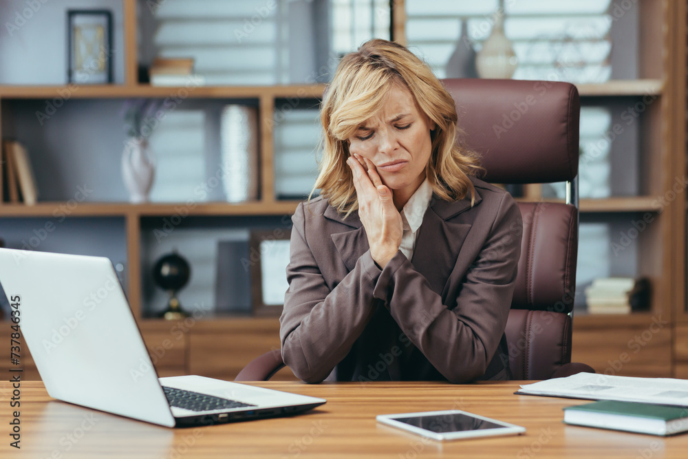 Mature businesswoman in pain, holding jaw suffering from toothache at office, showcasing workplace health issues. Mature businesswoman in pain, holding jaw suffering from toothache at office, showcasing workplace health issues.