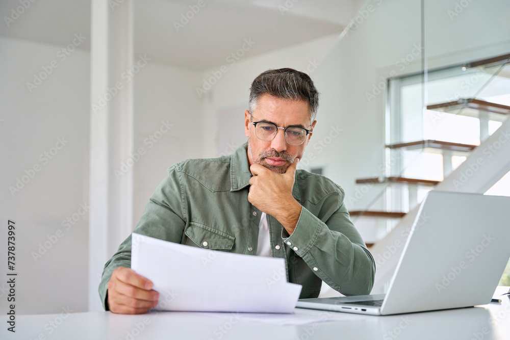 Serious middle aged mature man entrepreneur wearing eyeglasses looking at paper bill checking insurance invoice or tax document calculating expenses working at home using laptop computer. Serious middle aged mature man entrepreneur wearing eyeglasses looking at paper bill checking insurance invoice or tax document calculating expenses working at home using laptop computer.