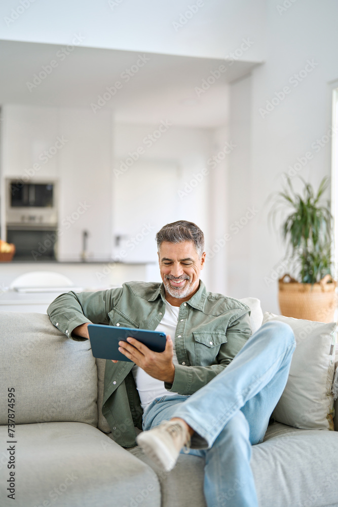 Smiling middle aged man using digital tablet relaxing on couch at home. Happy mature male user holding tab computer browsing internet on pad technology device sitting on sofa in living room. Vertical.