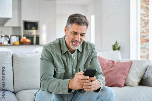 Middle aged old man using smartphone relaxing on couch at home. Happy senior mature male user holding cellphone browsing internet, texting messages on mobile cell phone technology sitting on sofa. photo