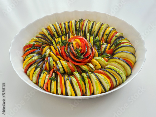 Ready-to-bake ratatouille in a deep dish on white background. Baking mold with zucchini, tomatoes, eggplant and herbs, sliced and beautifully arranged.