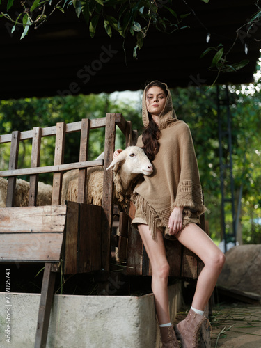 A woman in a brown sweater standing next to a sheep on a wooden bench in front of a tree