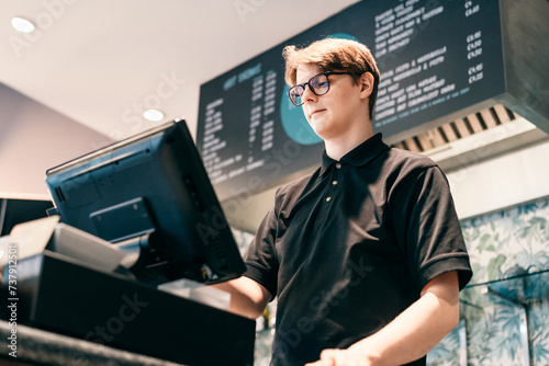 Young waiter serving customer at cash point in cafe. Man working with POS terminal. Cashier, barista checking for payment receipt. Hospitality, server and preparing a slip at the till in coffee shop photo