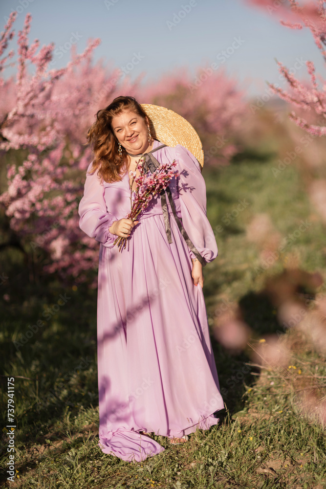 Woman blooming peach orchard. Against the backdrop of a picturesque peach orchard, a woman in a long pink dress and hat enjoys a peaceful walk in the park, surrounded by the beauty of nature.