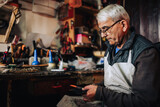 A senior craftsman is sitting at his workshop and cutting leather.