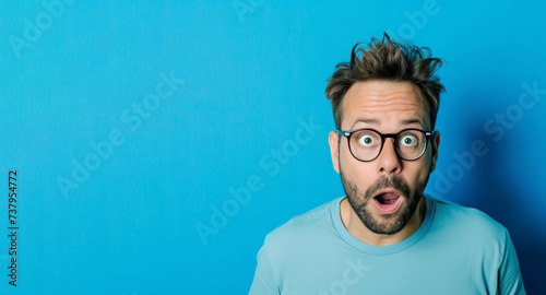surprised face of a young man in glasses on a blue background. The man opened his mouth in shock and surprise. Big eyes look into the camera. The surprise was a success. Place for text. Banner