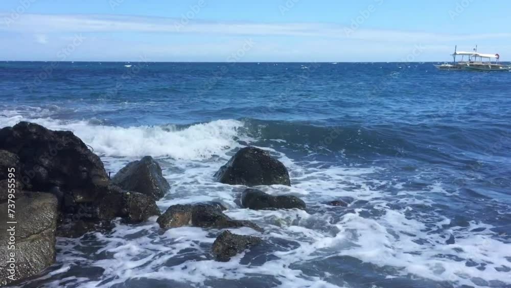 The waves crash in on the black rocks in the water. A boat is floating in the background—a sunny day in the Philippines—the beautiful view of the ocean in Malatapay.
