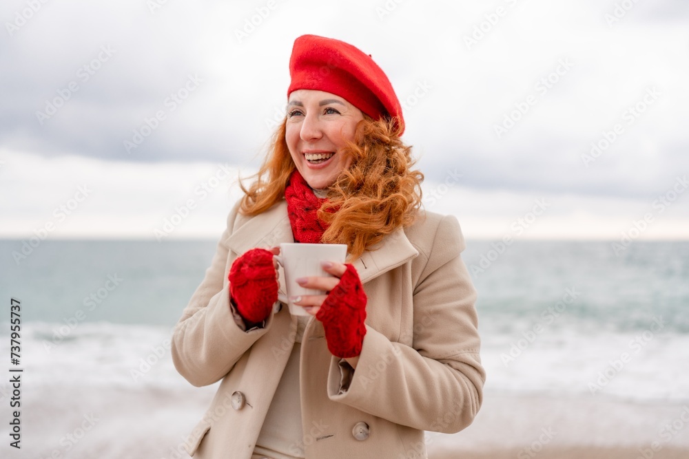 Obraz premium Woman beach in red beret scarf and mitts holding a cup of tea in his hands. Depicting beach relaxation and cozy attire. Walks by the sea