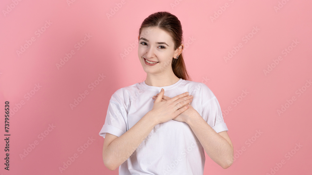 Fototapeta premium Portrait of a touched attractive Caucasian young brunette woman in a casual white t-shirt, touches the heart, the girl is grateful and says thank you, on a pink studio background.