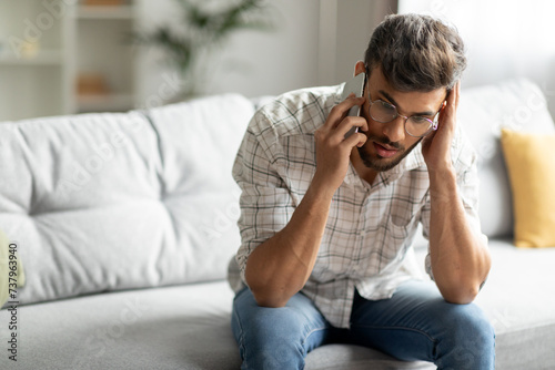 Confused indian guy talking on cellphone and touching head, having unpleasant mobile conversation at home, sitting on sofa in living room interior