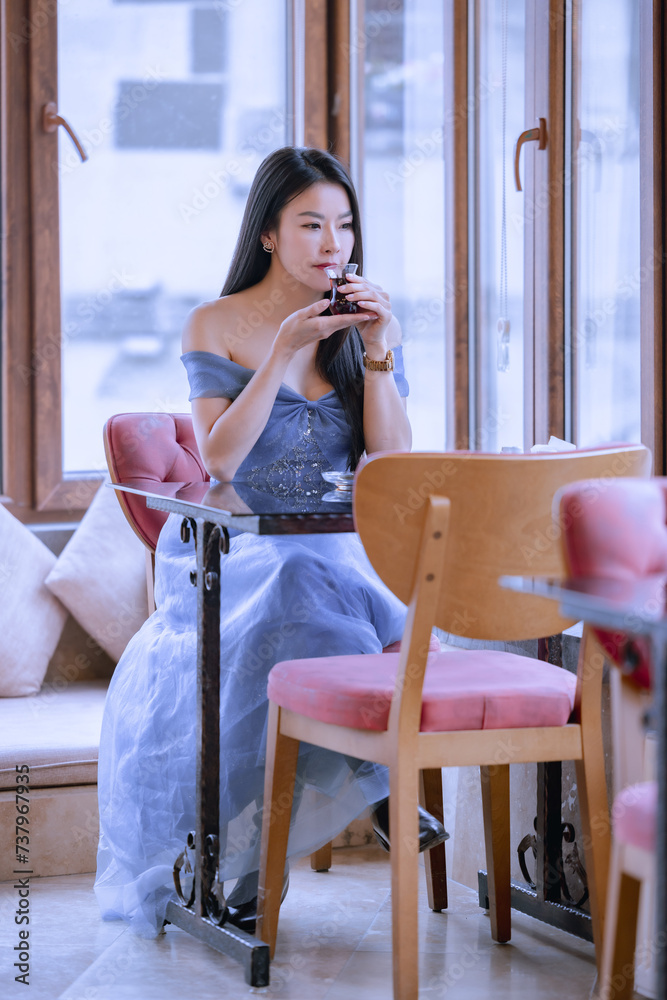 Beautiful princess in blue dress sitting and drinking a hot beverage while looking out of the window.Young Asian woman sipping hot tea at a table by the window.Relaxing time sipping hot tea or coffee. Beautiful princess in blue dress sitting and drinking a hot beverage while looking out of the window.Young Asian woman sipping hot tea at a table by the window.Relaxing time sipping hot tea or coffee.