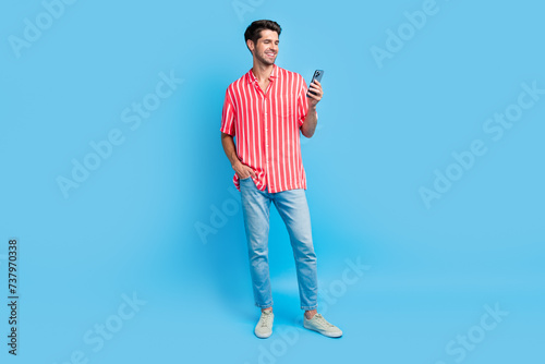 Full body photo of clever cheerful guy wear stylish shirt jeans trousers look at smartphone read email isolated on blue color background