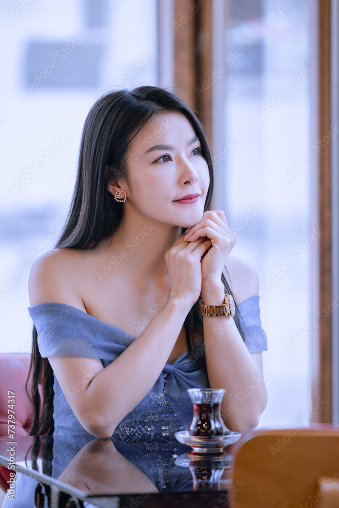 Beautiful princess in blue dress sitting and drinking a hot beverage while looking out of the window.Young Asian woman sipping hot tea at a table by the window.Relaxing time sipping hot tea or coffee. Beautiful princess in blue dress sitting and drinking a hot beverage while looking out of the window.Young Asian woman sipping hot tea at a table by the window.Relaxing time sipping hot tea or coffee.