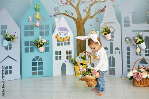 Baby girl celebrate Easter. Funny happy kid in bunny ears playing on Easter egg hunt. Family home decoration, colorful Easter eggs and flowers. Home decoration and flowers