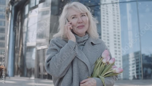 A senior woman talking by phone holding tulips outdoors