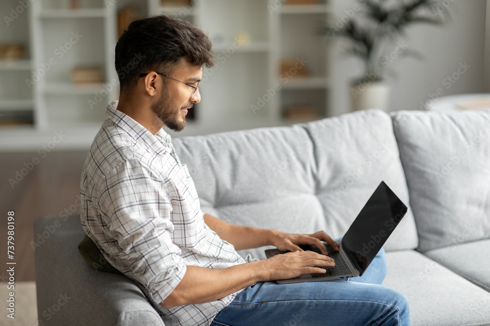 Young Indian man working online on laptop while sitting on couch at home, male freelancer browsing internet on computer, enjoying remote work Young Indian man working online on laptop while sitting on couch at home, male freelancer browsing internet on computer, enjoying remote work