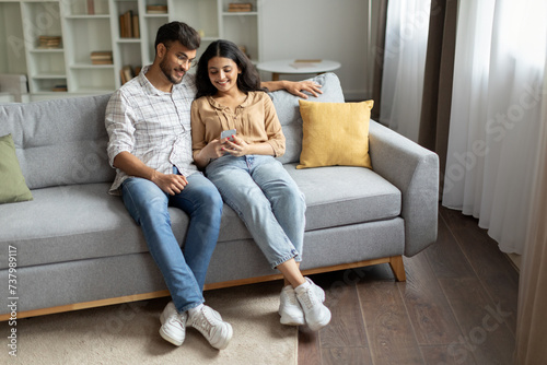 Indian couple using cellphone together, sitting in casual home interior on sofa, surfing internet or chatting with friends. Modern, digitally connected lifestyle