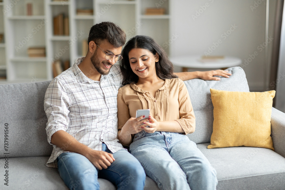 Happy indian spouses using mobile phone while relaxing on sofa, scrolling social media app or browsing internet, living room interior, free space