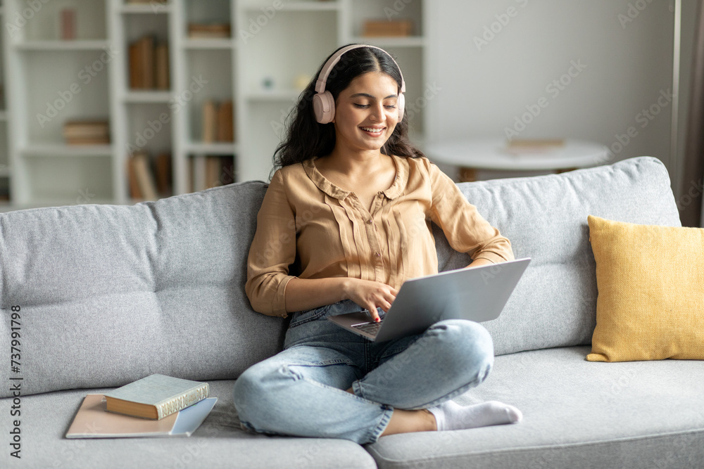 Indian lady wearing headphones and using laptop, enjoying digital leisure and e-learning, sitting on sofa at home interior, copy space Indian lady wearing headphones and using laptop, enjoying digital leisure and e-learning, sitting on sofa at home interior, copy space
