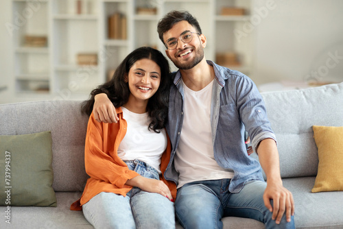 Loving indian spouses embracing, sitting on sofa in modern living room, husband hugging his wife and beaming with smiles together at camera