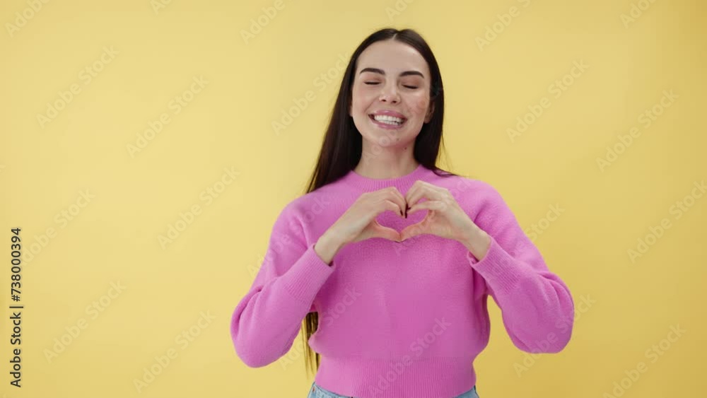 Portrait of cheerful caucasian lady making love sign with fingers and dance moves with body over bright yellow background. Happy brunette female dressed in pink sweater having romantic mood in studio.