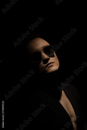 The face of a young man in glasses emerges from the shadows in a ray of light against a dark background