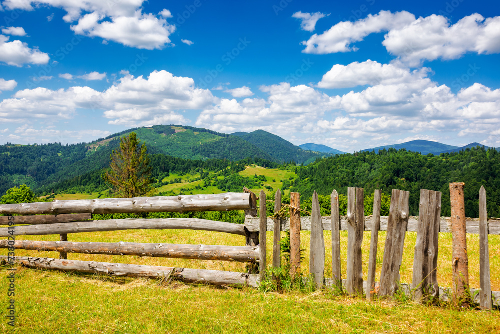 wooden fence on the meadow. mountainous rural landscape of transcarpathia, ukraine in summer. carpathian countryside with forested rolling hill beneath a blue sky with white fluffy clouds at high noon