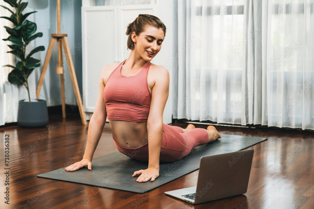 Fototapeta premium Flexible and dexterity woman in sportswear doing yoga position in meditation posture on exercising mat at home. Healthy gaiety home yoga online training session with peaceful mind and serenity.