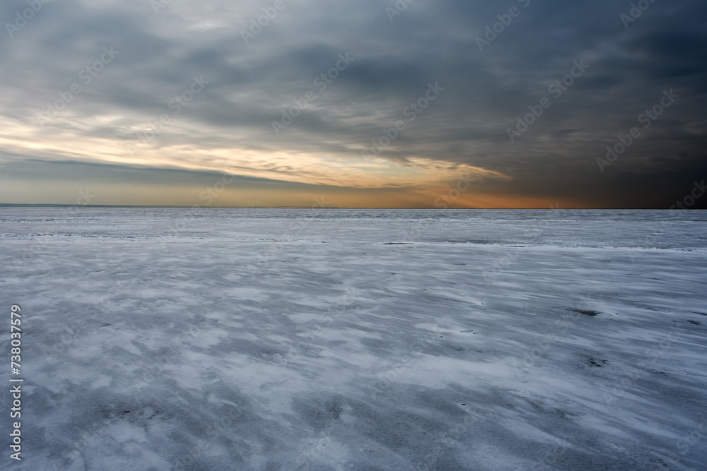 A frozen sea covered with dark ice and a thin layer of snow. Cloudy sky and orange sunlight at the beginning of sunset. The outlines of an industrial zone and a port are visible on the horizon