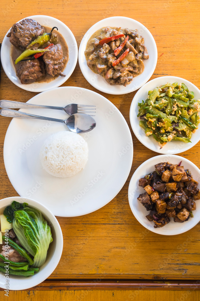 Top view of various Filipino cuisine dishes with steamed rice on a rustic wooden table, ideal for food blogs and menus.