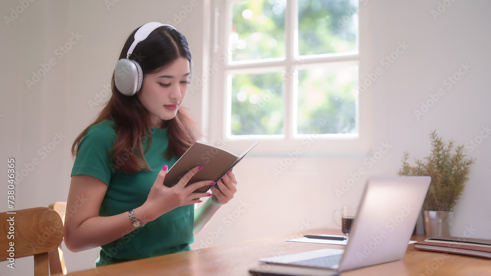 Young asian student women wearing headphone to studying and watching lecture webinar in online class on laptop while reading a book and learning knowledge remote education from home in living room