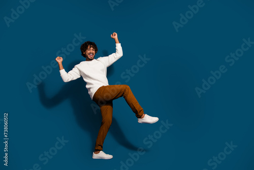 Full length photo of excited lucky guy dressed white pullover rising fists walking empty space isolated dark blue color background