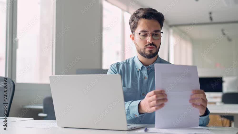 Busy serious young business man working on laptop computer holding documents at workplace. Latin professional businessman employee or manager thinking of financial management in office.