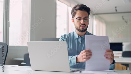 Busy serious young business man working on laptop computer holding documents at workplace. Latin professional businessman employee or manager thinking of financial management in office. photo