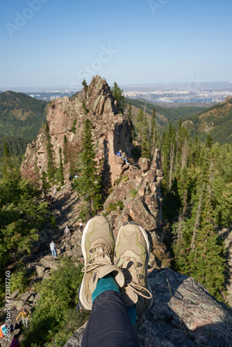 Reach the top of cliff. Legs of a man in hiking boots against the background of igneous rocks and green forest near the city. Danger of sitting on the edge of rock without insurance. Amazing nature