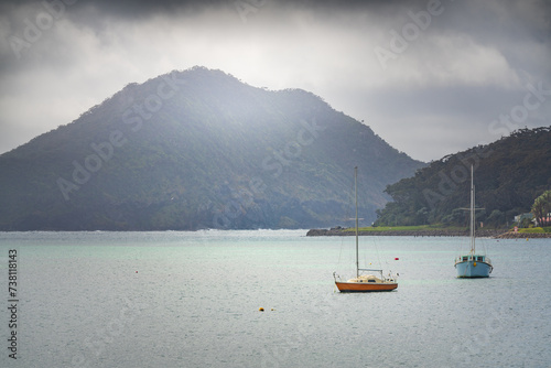 Two small yachts on a calm bay beneath a high coastal mountain with a dark sky building above photo