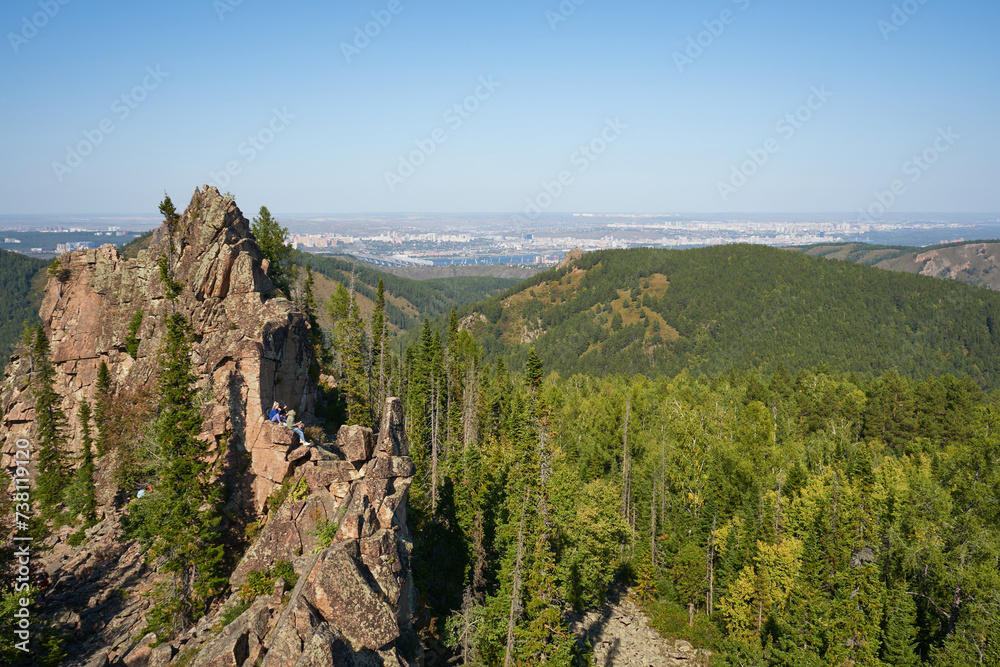 Group of tourists sits on an igneous rock against the backdrop of green forest and city in the distance. Summer natural landscape. Active lifestyle: climbing, hiking and tourism. Enjoying nature