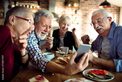 Senior friends laughing and sharing photos on smartphone at breakfast