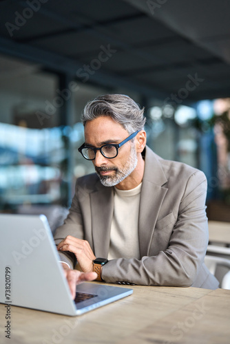 Busy older business man hybrid working outdoors using laptop. Middle aged business man executive wearing suit and glasses sitting outside office looking at computer browsing web, vertical.
