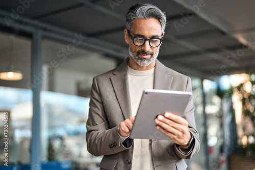 Busy mature older business man executive standing in office using digital tablet. Middle aged professional businessman corporate manager wearing suit and glasses holding tab working on finance project