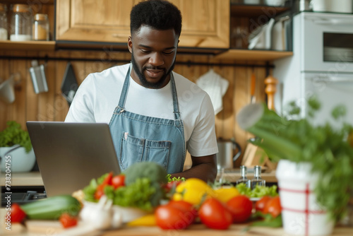 Portrait of young African American man preparing fresh vegetable salad looking at recipes on a laptop standing in the kitchen at home