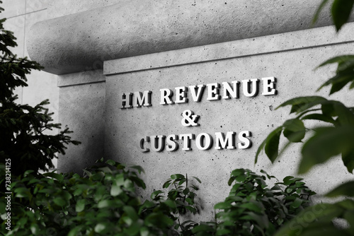 Silver sign of Home Office Revenue and Customs on a concrete wall with green plants as foreground. Illustration of the concept of taxation