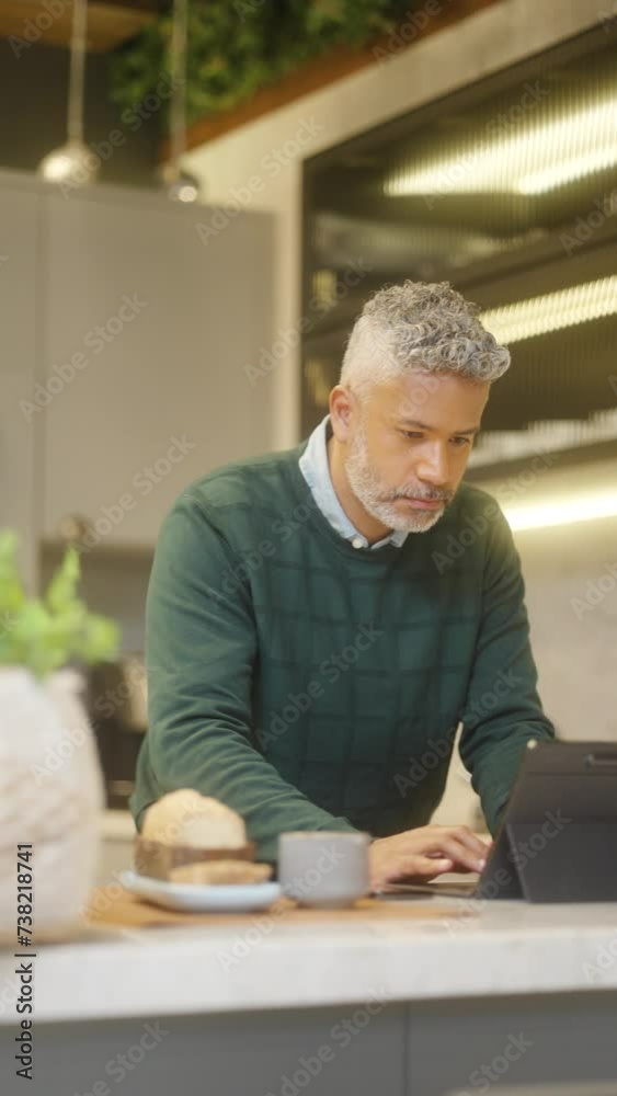 Focused Home Workspace: Silver-Haired Man in Casual Sweater Engaging in Remote Business Using Digital Tablet in Stylish Kitchen. Focused Home Workspace: Silver-Haired Man in Casual Sweater Engaging in Remote Business Using Digital Tablet in Stylish Kitchen.
